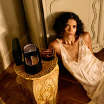 Woman in a white dress sitting next to a decorative stool with a suitcase on a wooden floor.