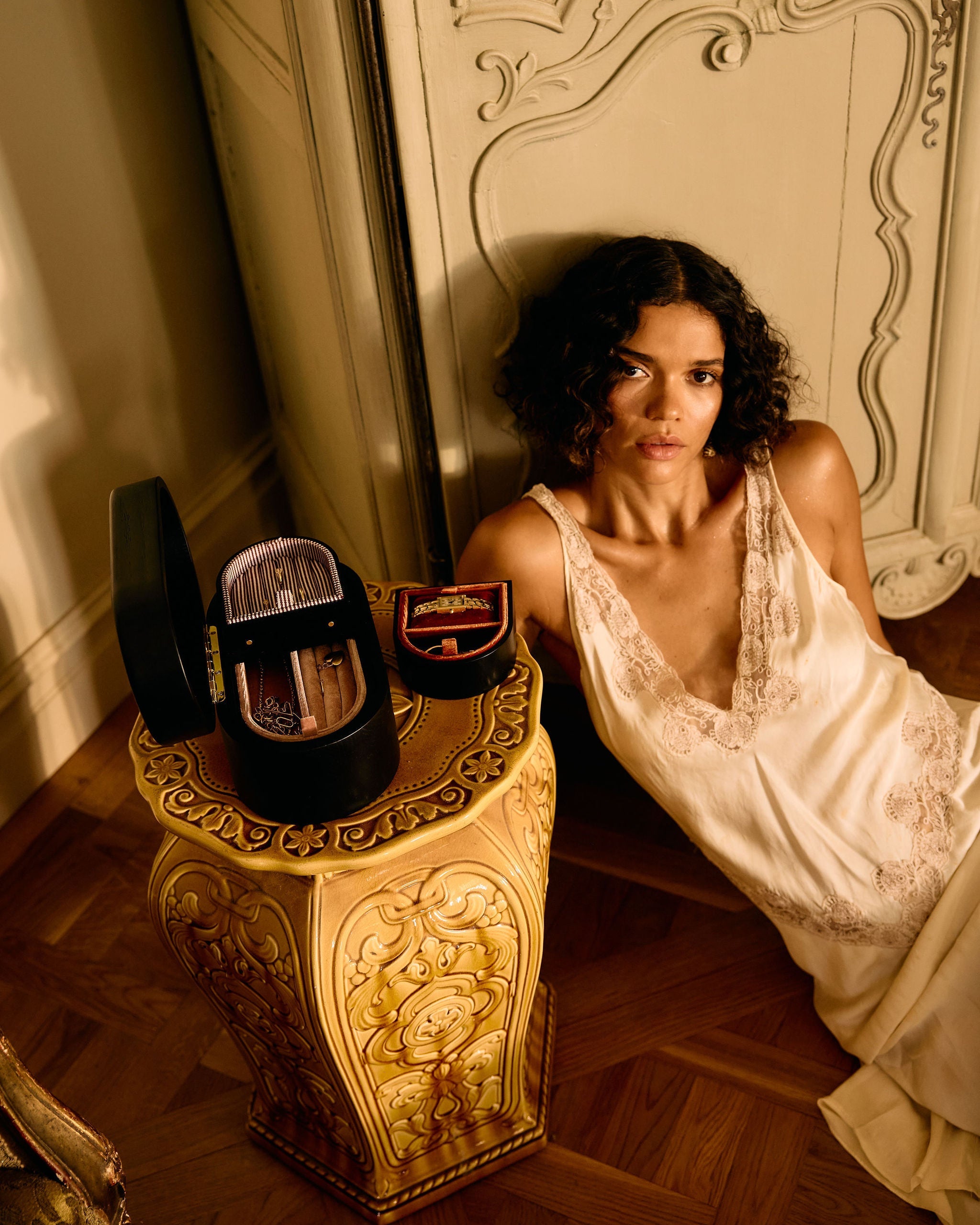 Woman in a white dress sitting next to a decorative stool with a suitcase on a wooden floor.