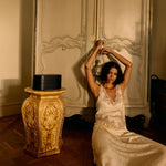 Woman in a white dress sitting in front of an ornate wardrobe in a dimly lit room.