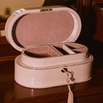 A light pink oval jewelry box with an open lid, showing interior compartments and a tassel charm, placed on a dark wooden surface in a warm indoor setting.