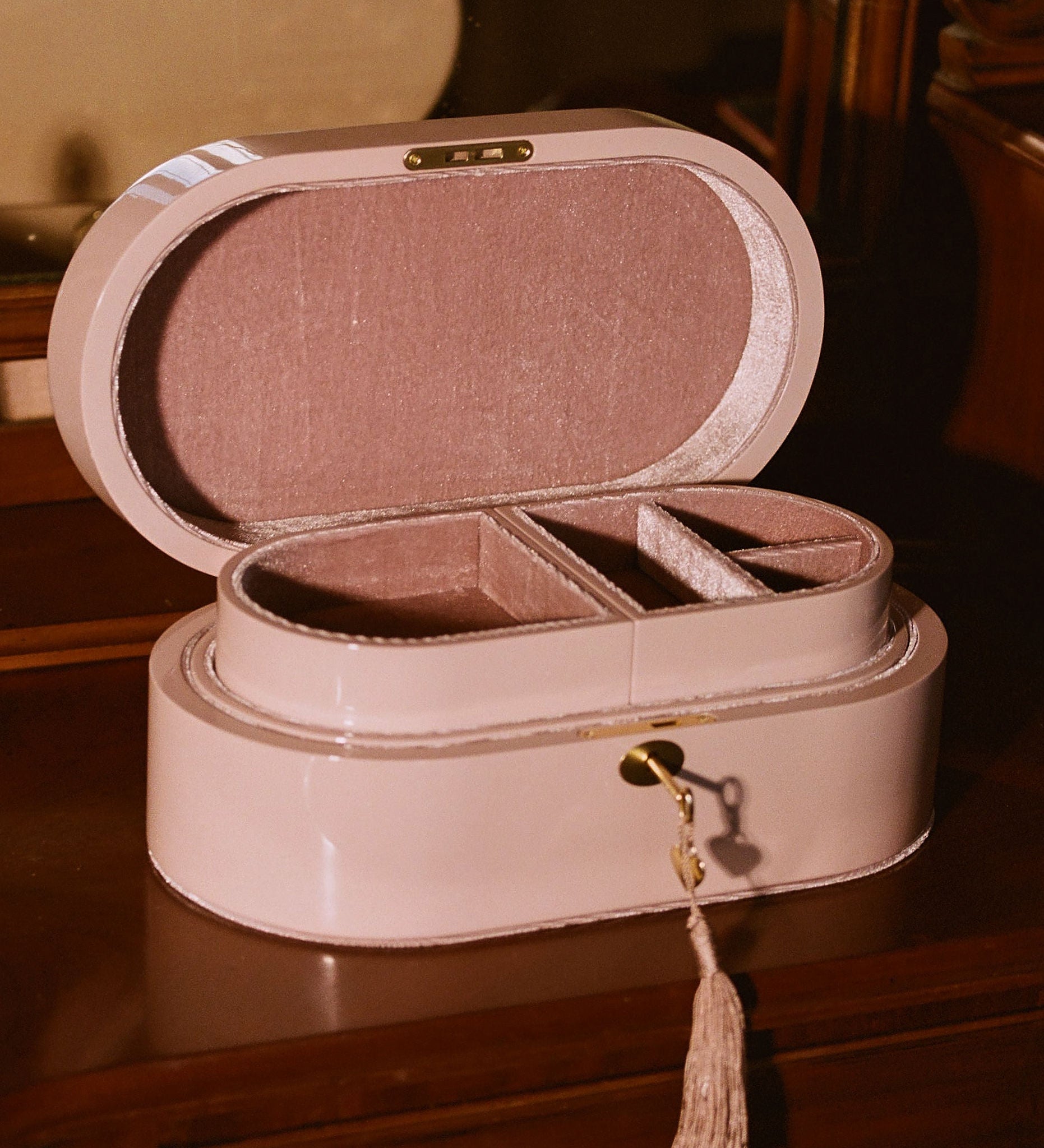 A light pink oval jewelry box with an open lid, showing interior compartments and a tassel charm, placed on a dark wooden surface in a warm indoor setting.