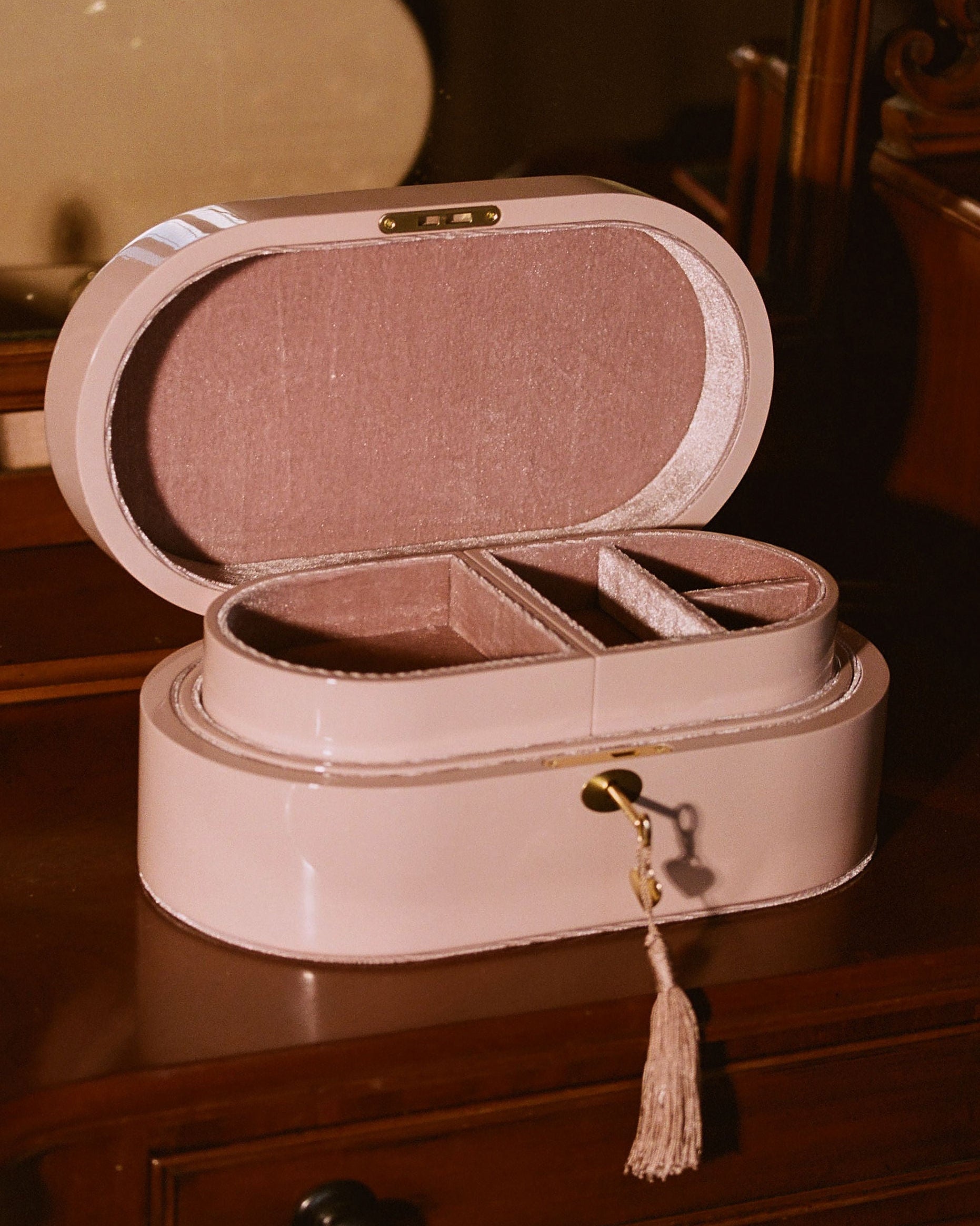 A light pink oval jewelry box with an open lid, showing interior compartments and a tassel charm, placed on a dark wooden surface in a warm indoor setting.