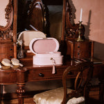 A vintage pink makeup case with an open lid sits on an ornate dark wood vanity table with a matching chair, set against a warm pink wall.