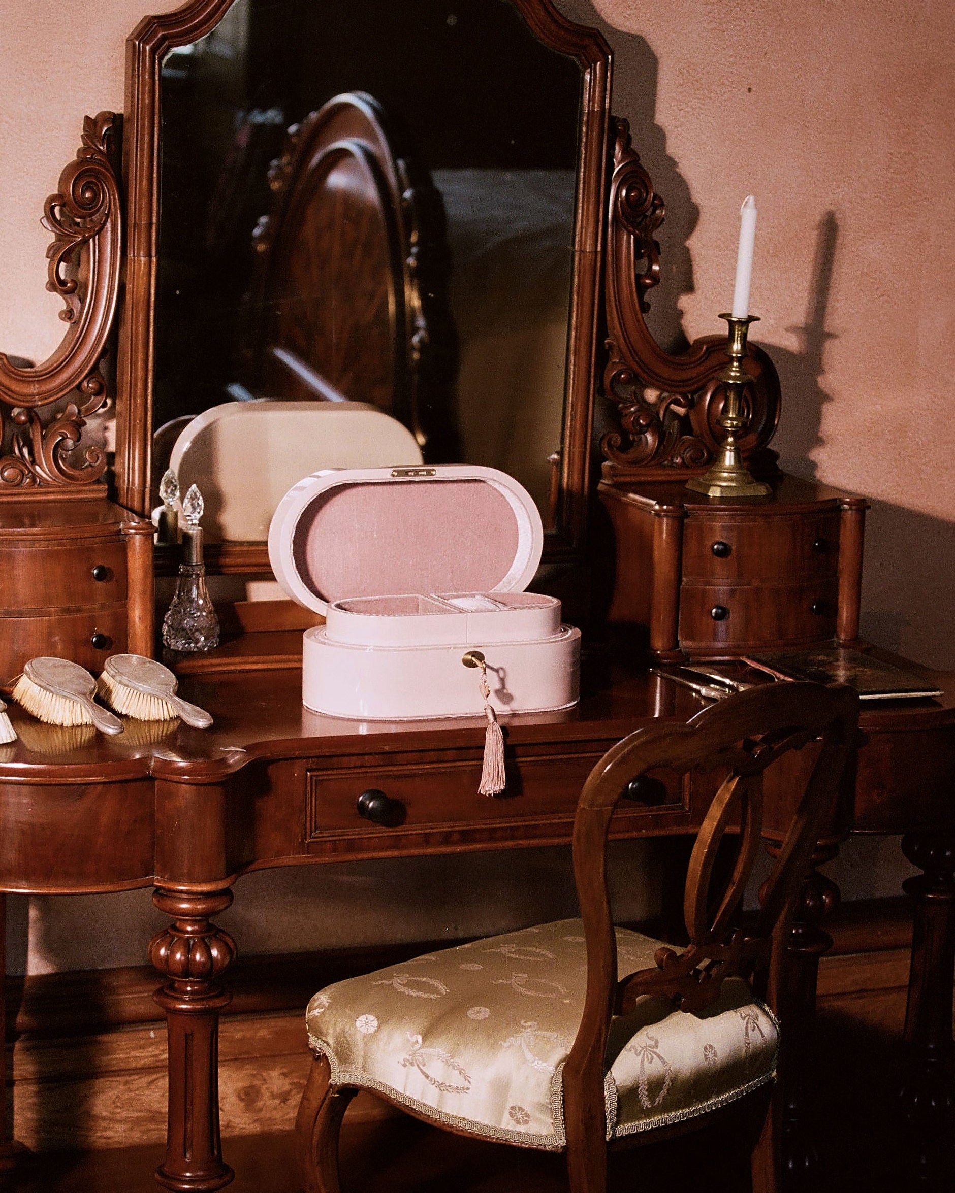 A vintage pink makeup case with an open lid sits on an ornate dark wood vanity table with a matching chair, set against a warm pink wall.
