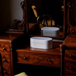 A vintage dark wood vanity table with an attached mirror, featuring carved details and multiple drawers, with a white oval cosmetic box placed on top.