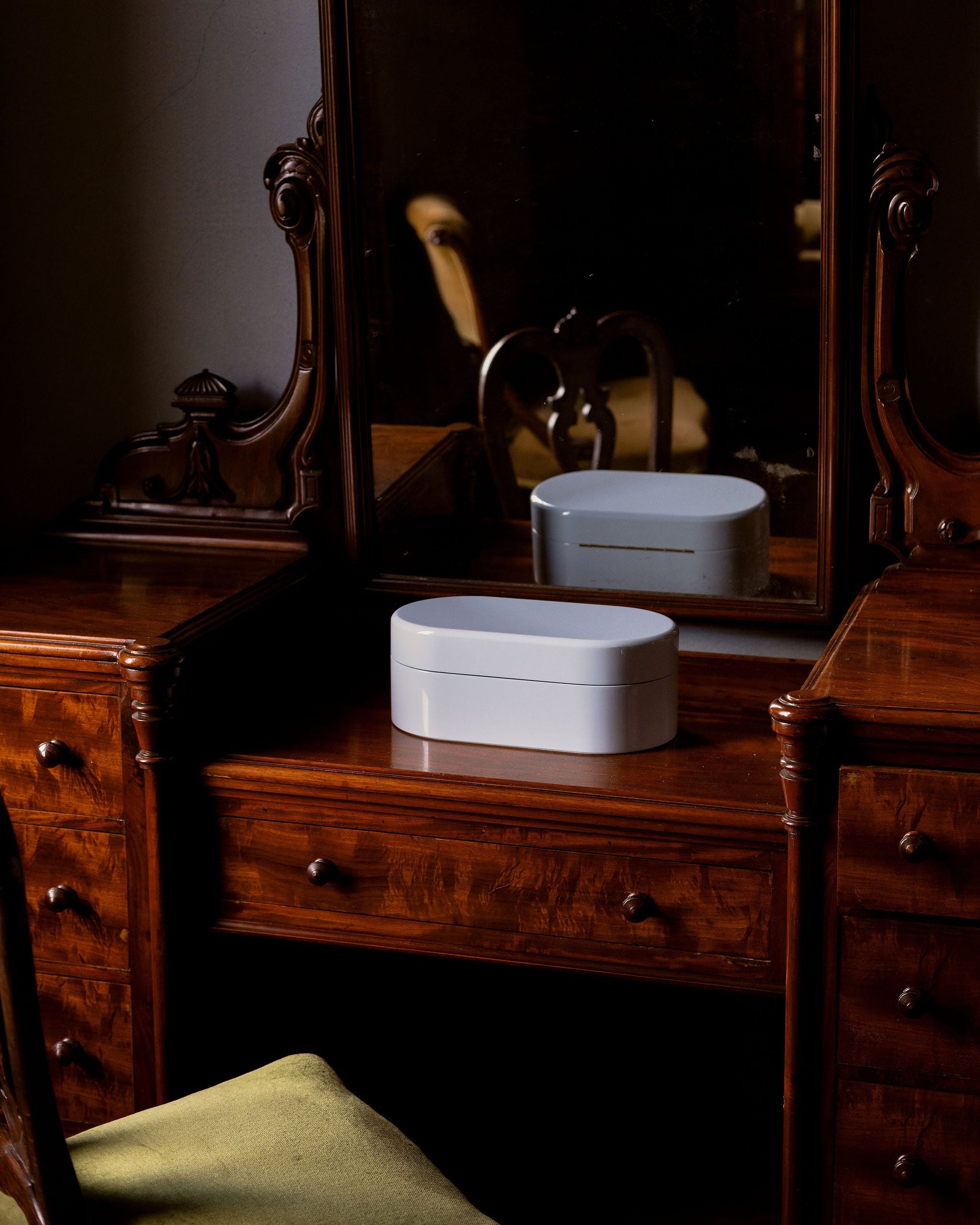 A vintage dark wood vanity table with an attached mirror, featuring carved details and multiple drawers, with a white oval cosmetic box placed on top.