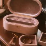 A blush pink velvet jewelry box set displayed on a dark wooden vanity table. The set includes a large oval-shaped box with its lid open, showing a velvet-lined interior with a divider, a smaller matching square tray holding a gold ring, and a small tassel accessory.