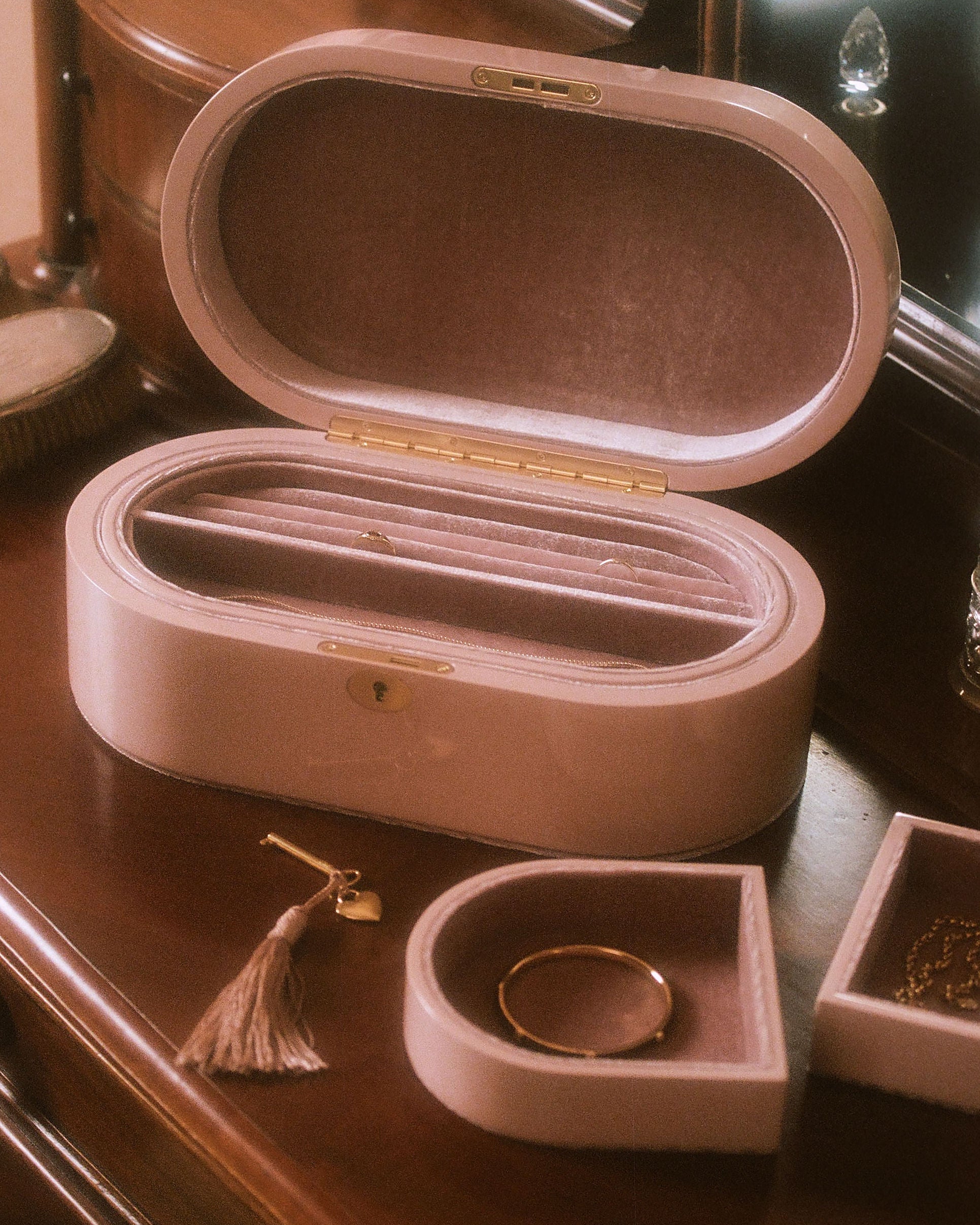 A blush pink velvet jewelry box set displayed on a dark wooden vanity table. The set includes a large oval-shaped box with its lid open, showing a velvet-lined interior with a divider, a smaller matching square tray holding a gold ring, and a small tassel accessory.