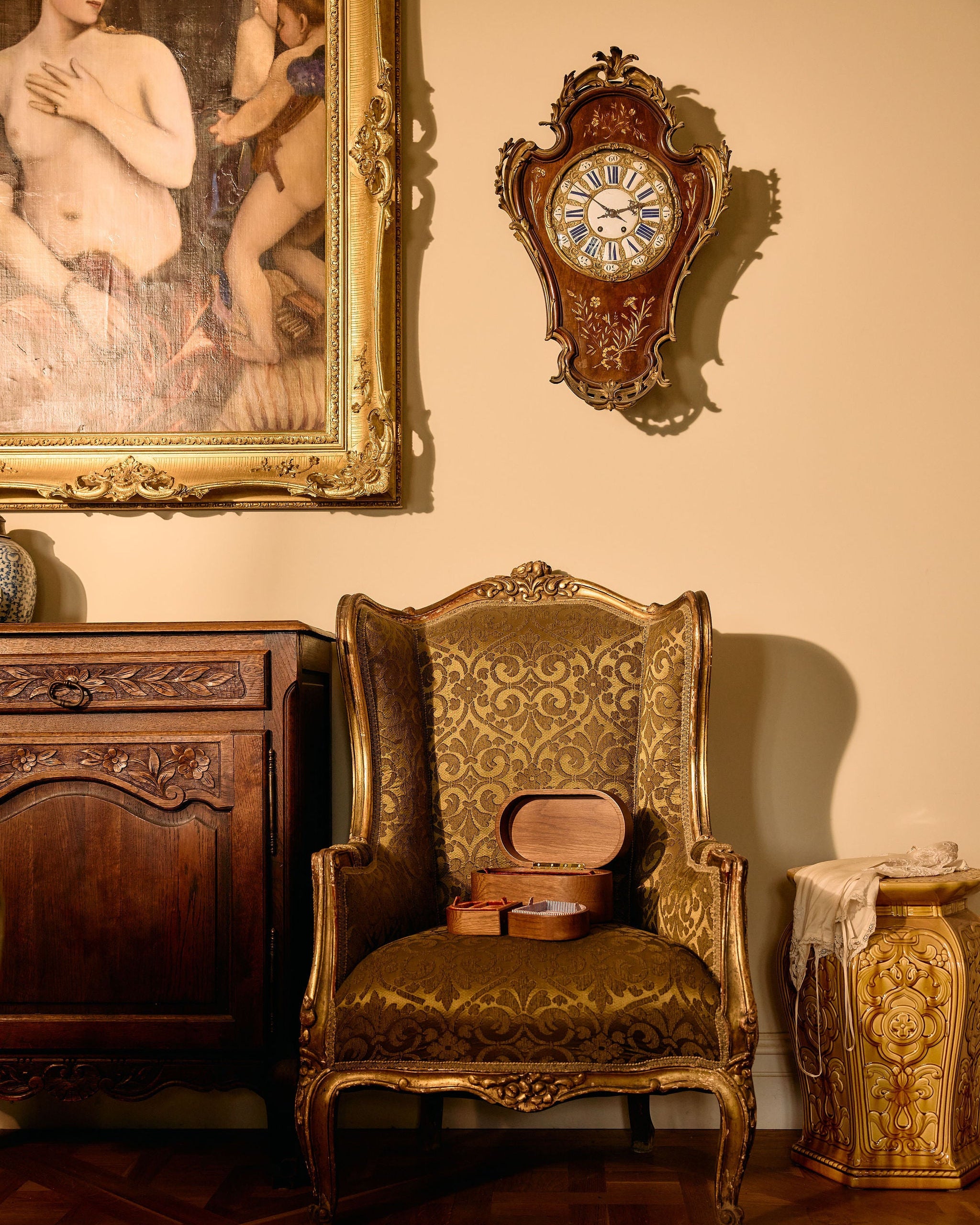 Vintage room with ornate chair, clock, and painting on a beige wall.