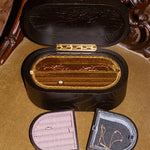 A vintage black wooden ring box with a gold-tone hinge and interior, shown open with two smaller ring boxes placed in front on a tan upholstered chair.