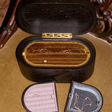 A vintage black wooden ring box with a gold-tone hinge and interior, shown open with two smaller ring boxes placed in front on a tan upholstered chair.