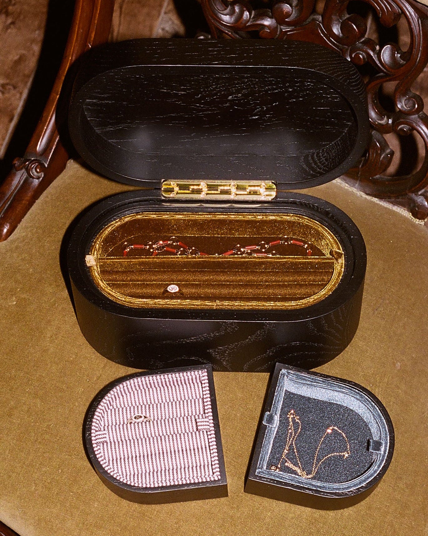 A vintage black wooden ring box with a gold-tone hinge and interior, shown open with two smaller ring boxes placed in front on a tan upholstered chair.