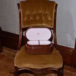 A vintage brown wooden box with an open lid resting on an antique upholstered chair with brown velvet fabric, set against a gray wall and wooden floor.