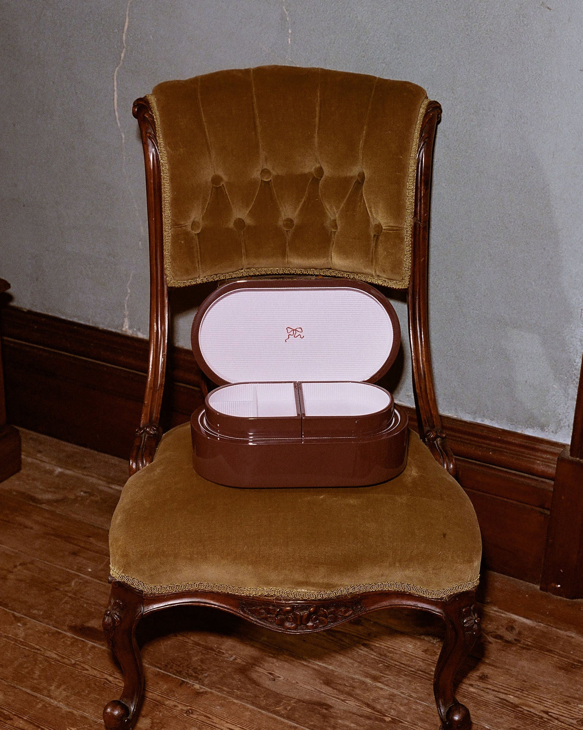 A vintage brown wooden box with an open lid resting on an antique upholstered chair with brown velvet fabric, set against a gray wall and wooden floor.