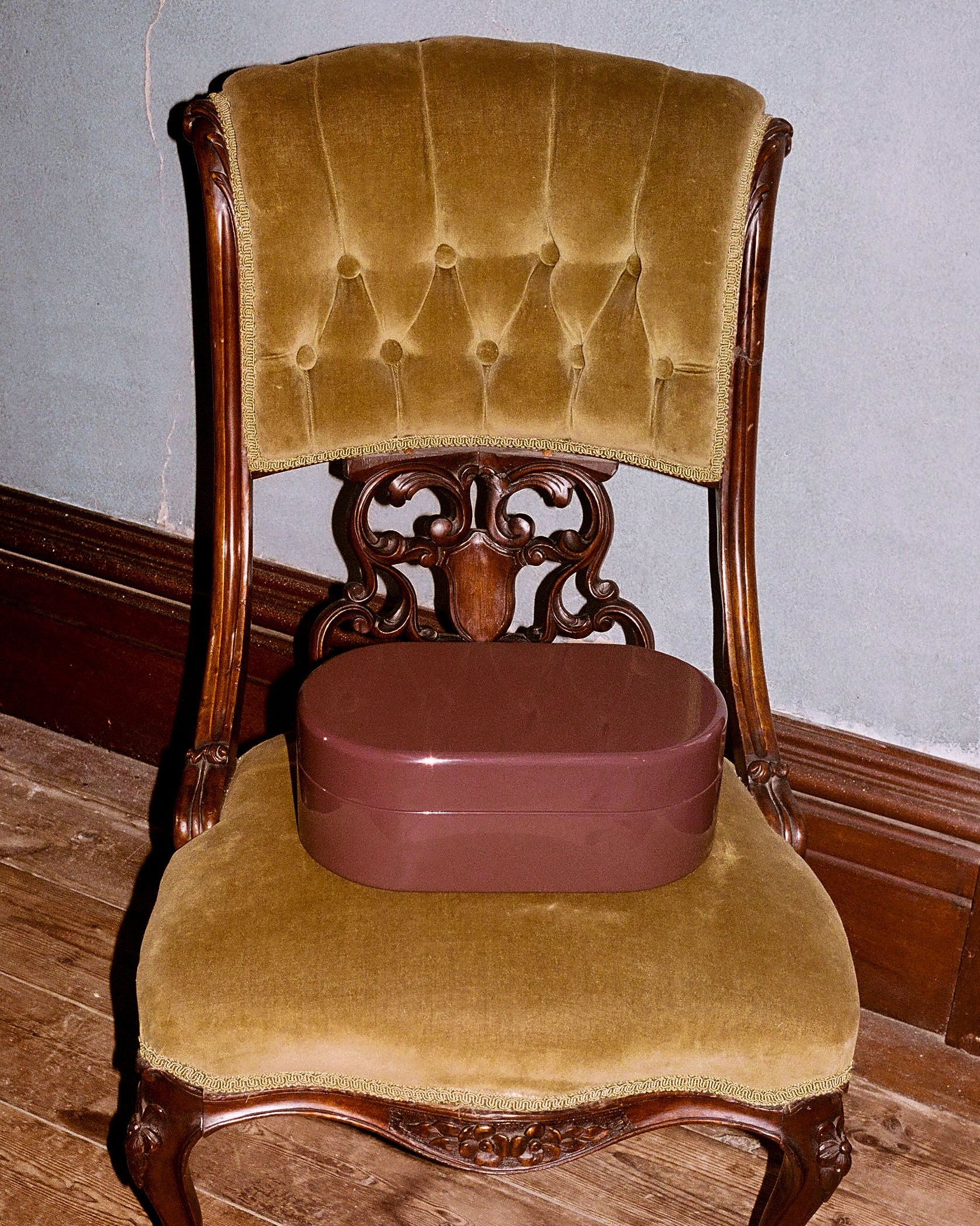 An antique carved wooden chair with mustard yellow velvet upholstery, featuring a round brown storage box on its seat, set against a gray wall and wooden floor.