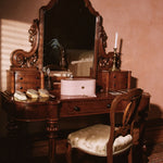 An ornate dark wood vanity table with a large arched mirror, matching side tables, a pink makeup case, and a cushioned wooden chair in a warmly lit room.