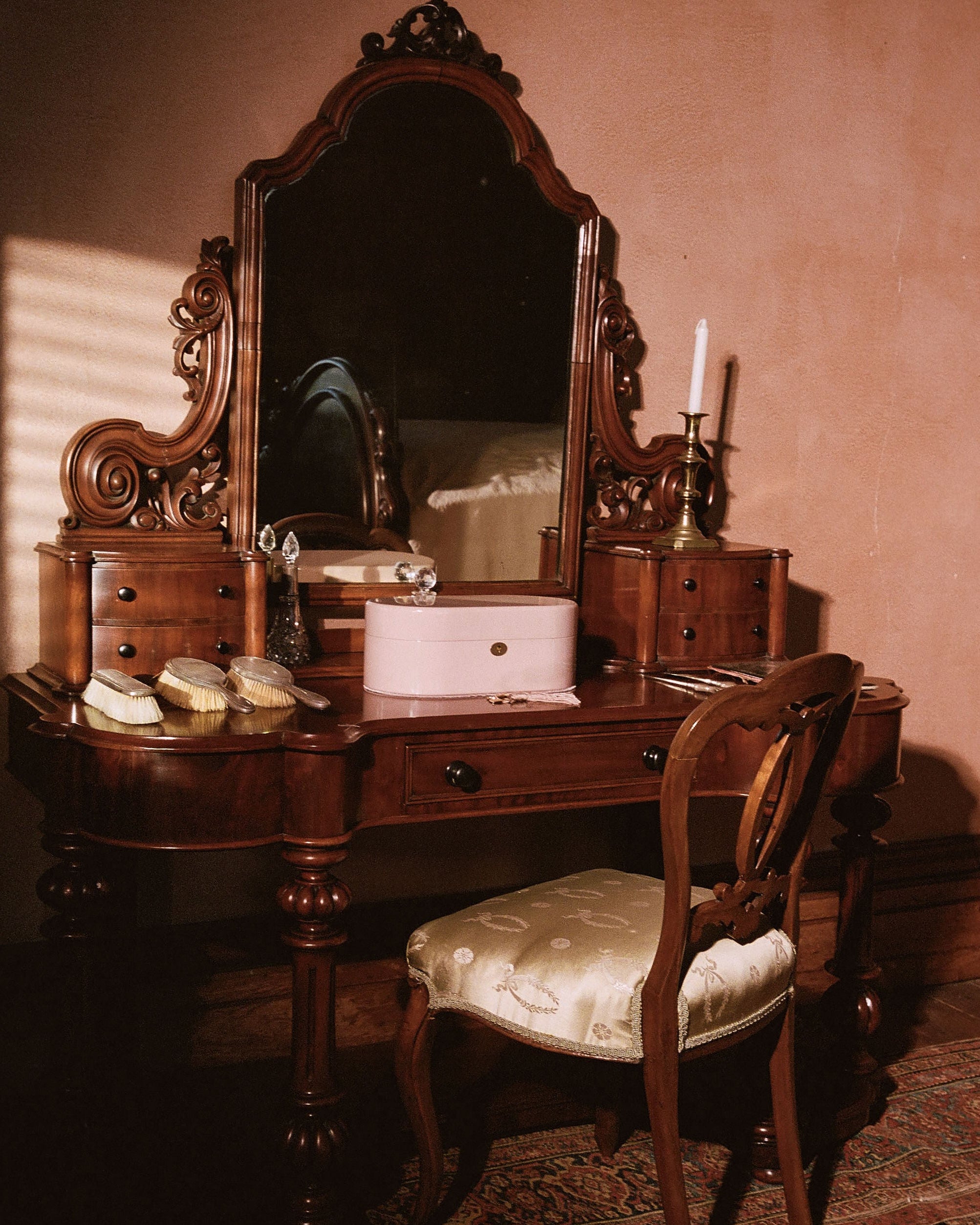 An ornate dark wood vanity table with a large arched mirror, matching side tables, a pink makeup case, and a cushioned wooden chair in a warmly lit room.