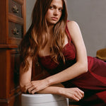 A woman with long brown hair wearing a dark red sleeveless dress kneels on a white stool in a dimly lit room next to a wooden chest of drawers.