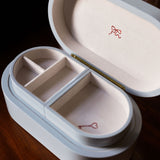 A white oval jewelry box with an open lid, featuring two interior compartments and small red ribbon and key motifs printed inside, set on a dark wooden surface.