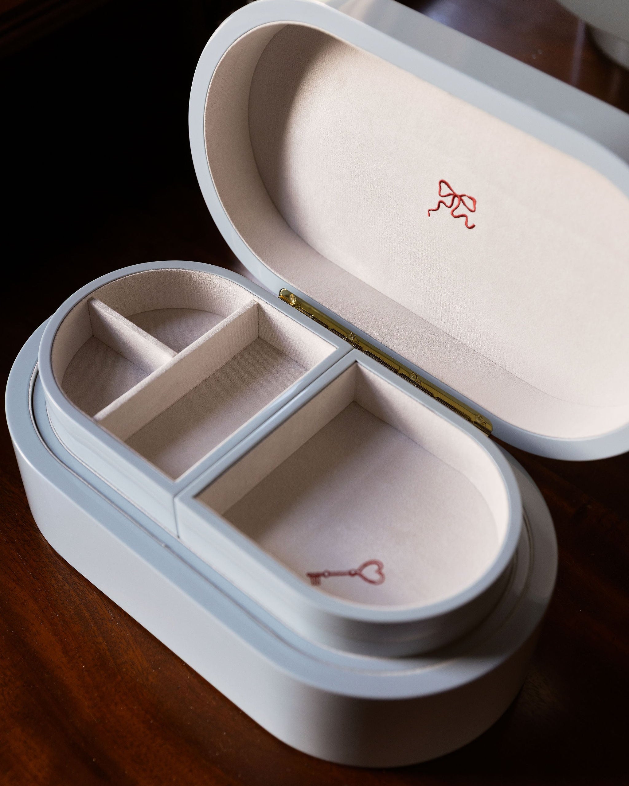 A white oval jewelry box with an open lid, featuring two interior compartments and small red ribbon and key motifs printed inside, set on a dark wooden surface.