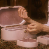 A person's hands open a round, pale pink jewelry box with its lid propped open, revealing a velvet-lined interior. A smaller matching heart-shaped jewelry box sits in the foreground on a patterned rug, with another box partially visible on the right.