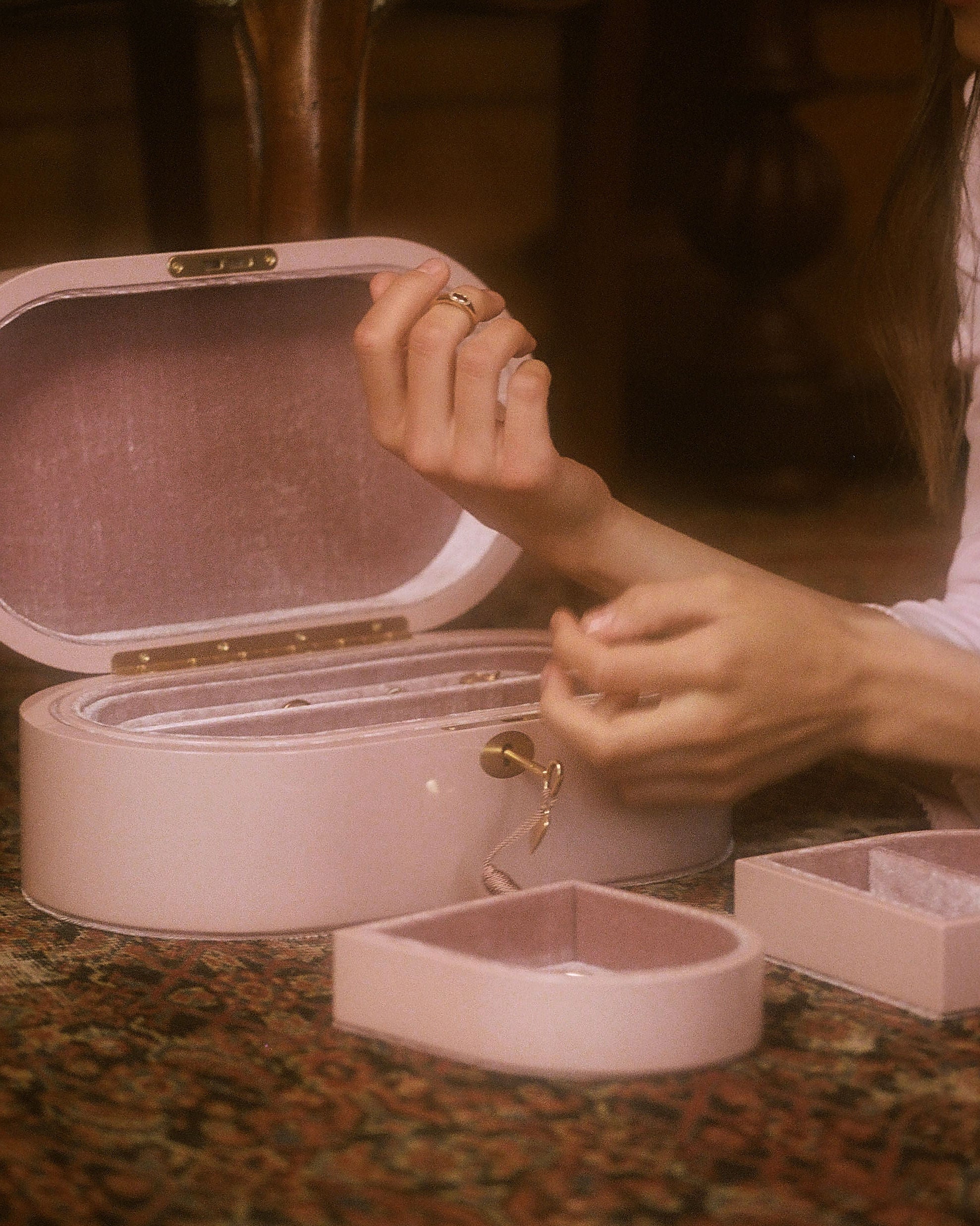 A person's hands open a round, pale pink jewelry box with its lid propped open, revealing a velvet-lined interior. A smaller matching heart-shaped jewelry box sits in the foreground on a patterned rug, with another box partially visible on the right.