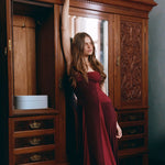A woman in a floor-length burgundy gown poses in front of a large carved wooden wardrobe in a dimly lit room.