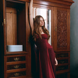 A woman in a floor-length burgundy gown poses in front of a large carved wooden wardrobe in a dimly lit room.