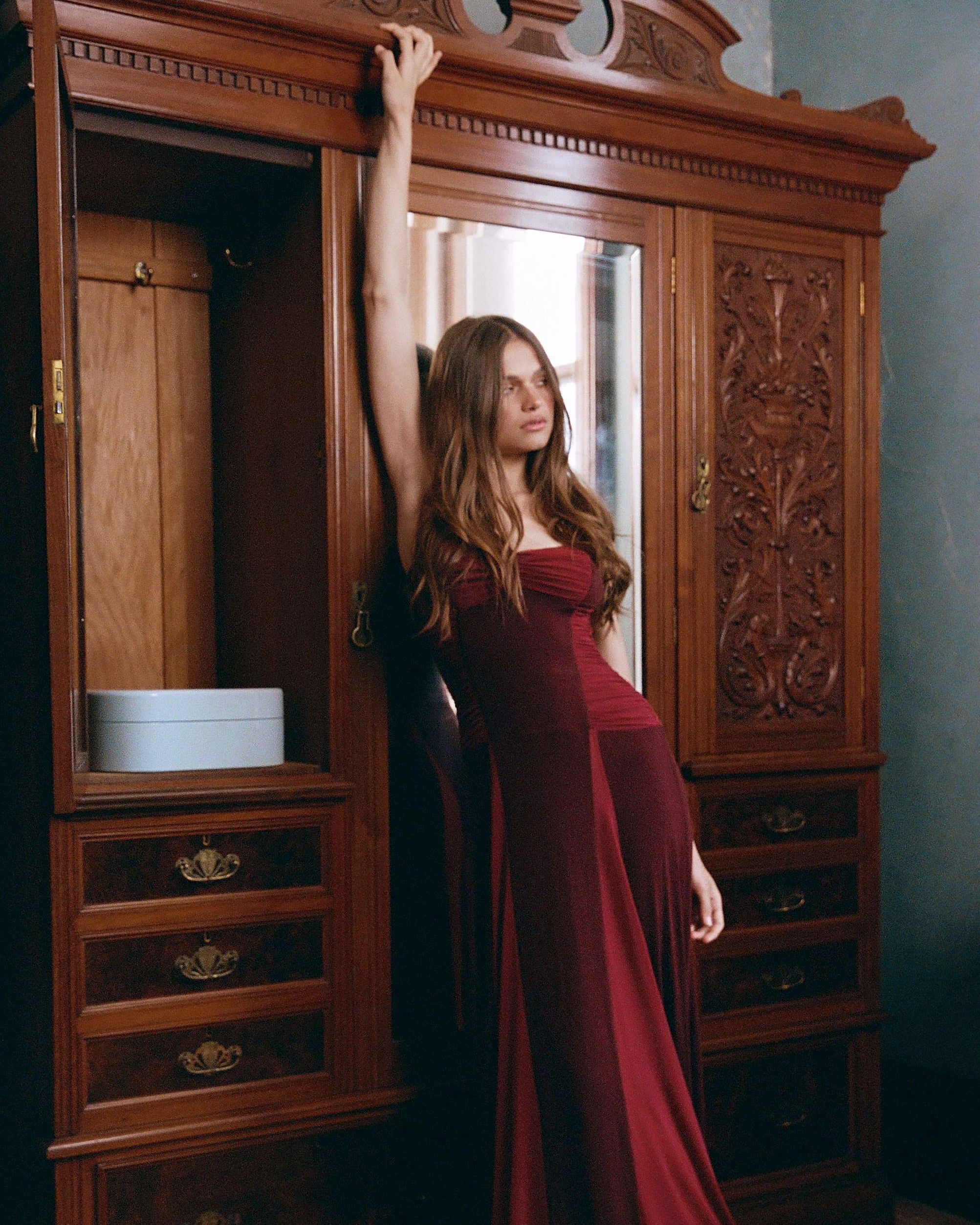 A woman in a floor-length burgundy gown poses in front of a large carved wooden wardrobe in a dimly lit room.